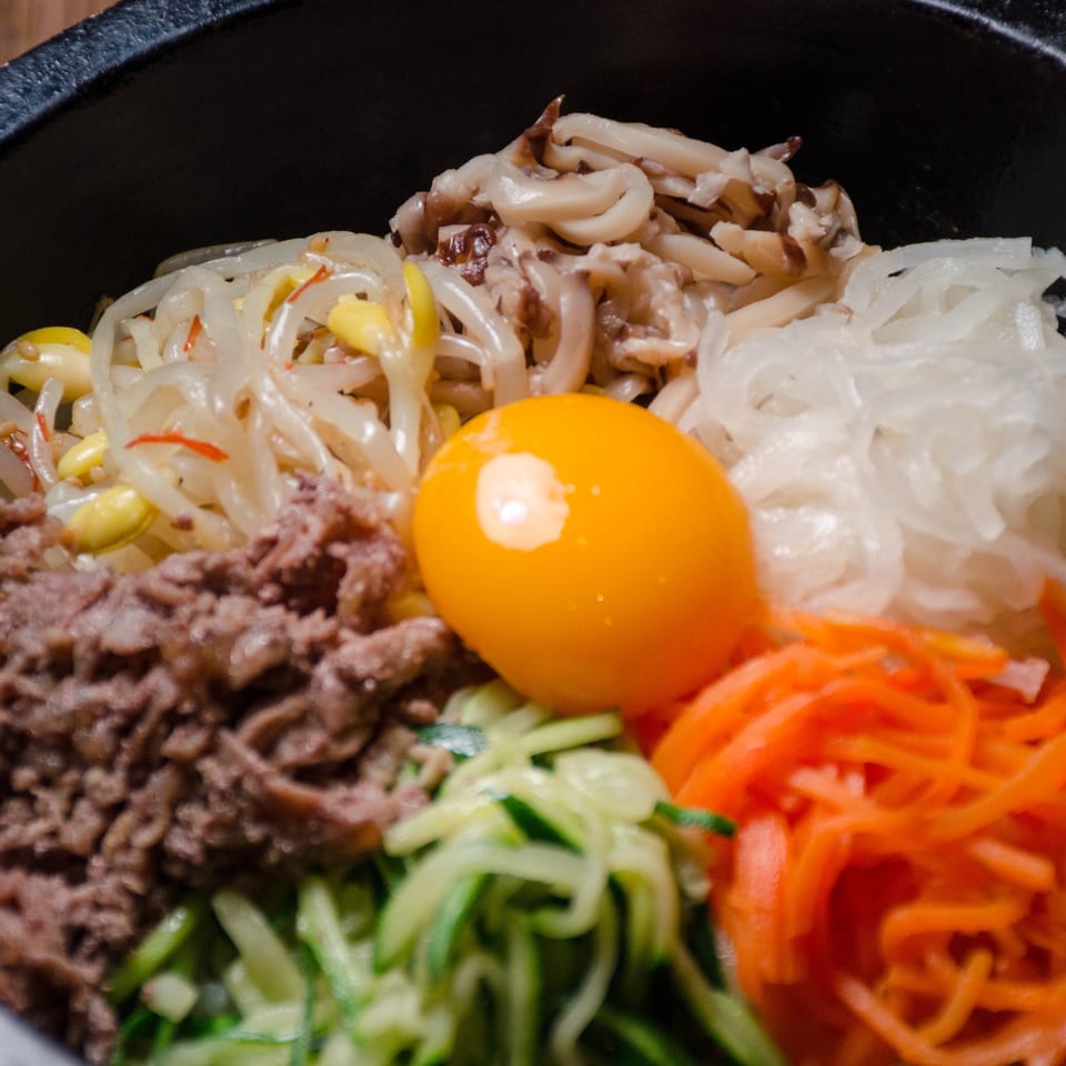 Close-up of a Korean bibimbap bowl with beef, vegetables, mushrooms, noodles, and a raw egg yolk.