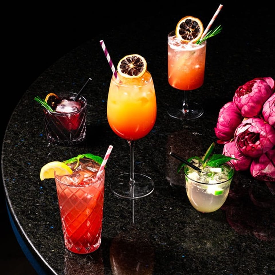 Five colorful cocktails with citrus garnishes and straws arranged on a glossy black table beside bright pink flowers.