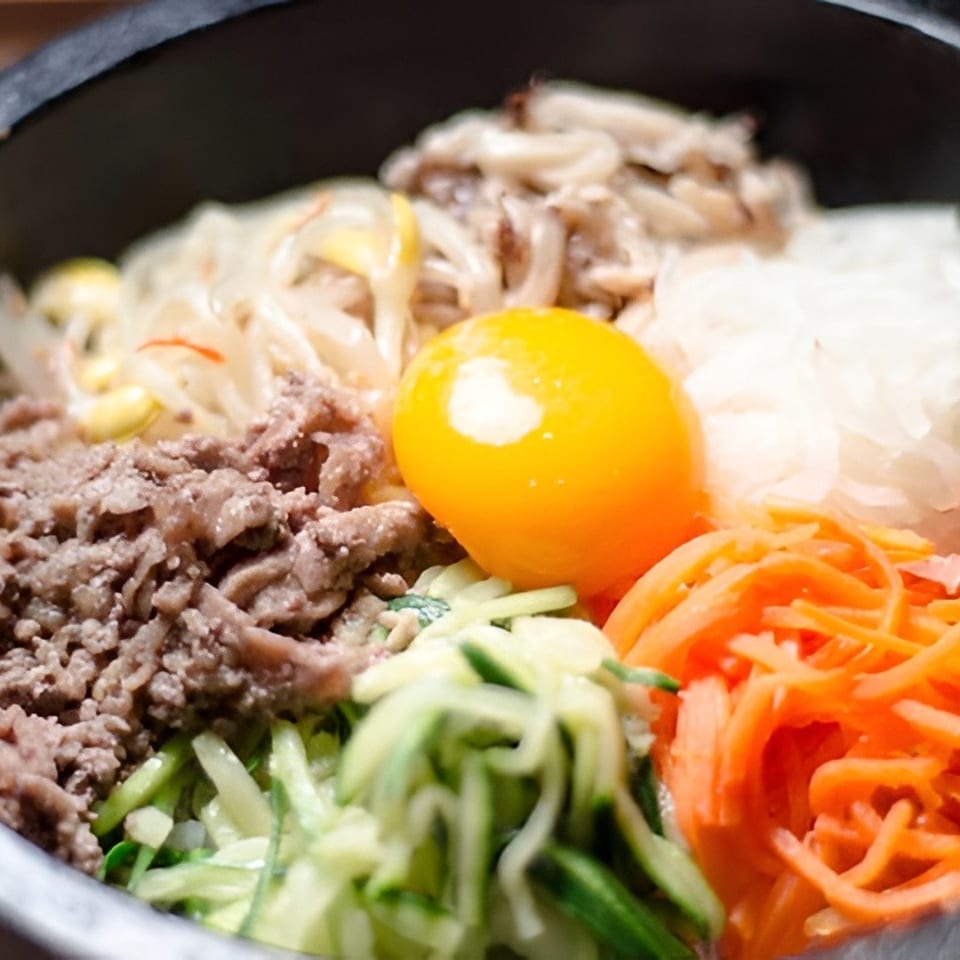 Korean bibimbap in a black bowl with beef, vegetables, mushrooms, shredded carrots, and a bright raw egg yolk.