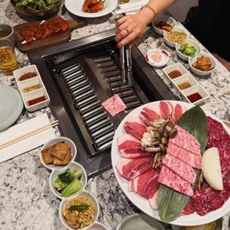 Korean barbecue table with marbled meats, vegetables, side dishes, and tongs grilling food on a tabletop stove.