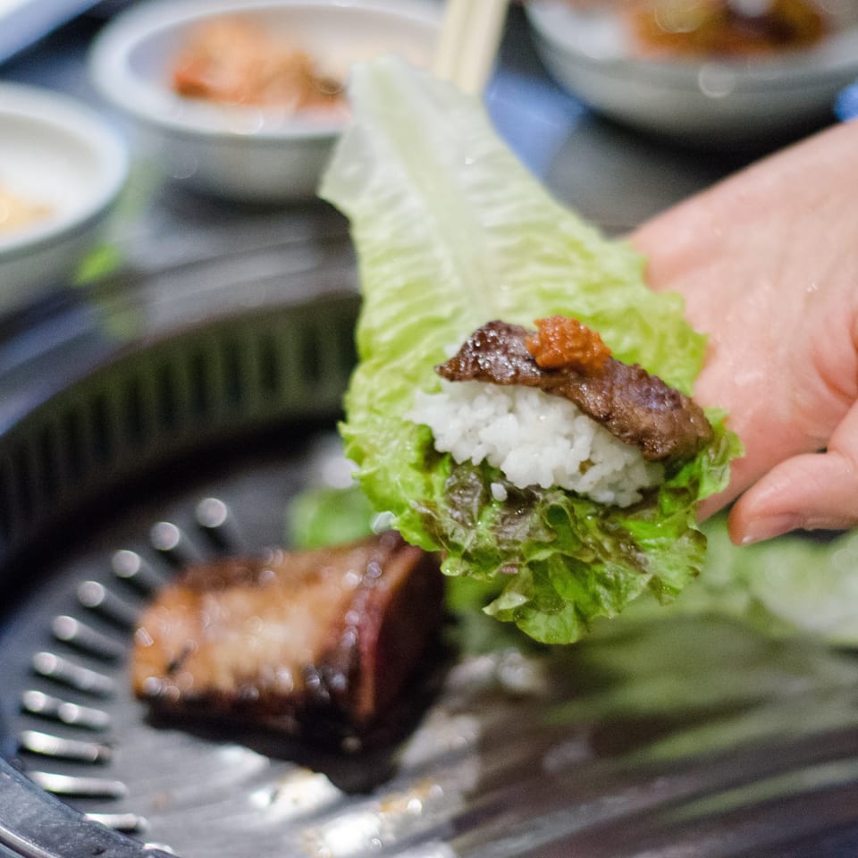 Hand assembles a lettuce wrap with white rice, grilled meat, and chili paste over a plate surrounded by side dishes.