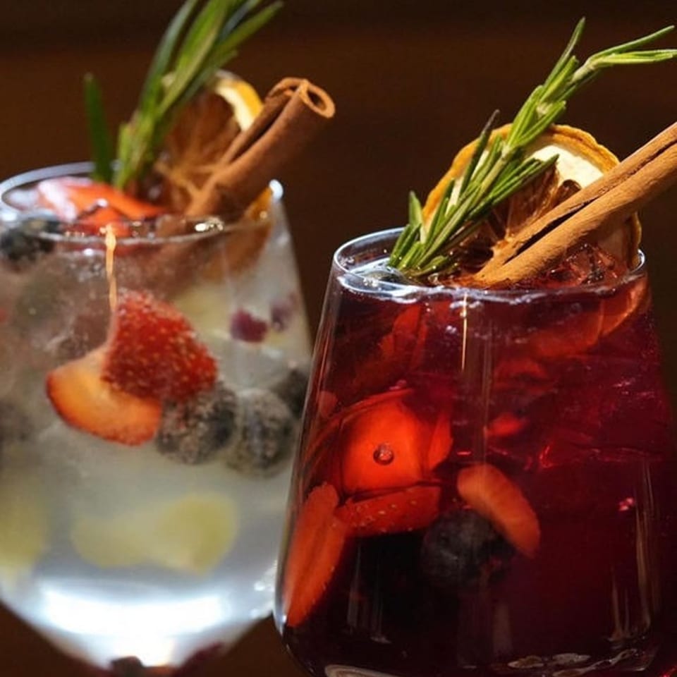 Two fruit-filled cocktails garnished with rosemary, cinnamon sticks, and citrus slices sit against a dark background.