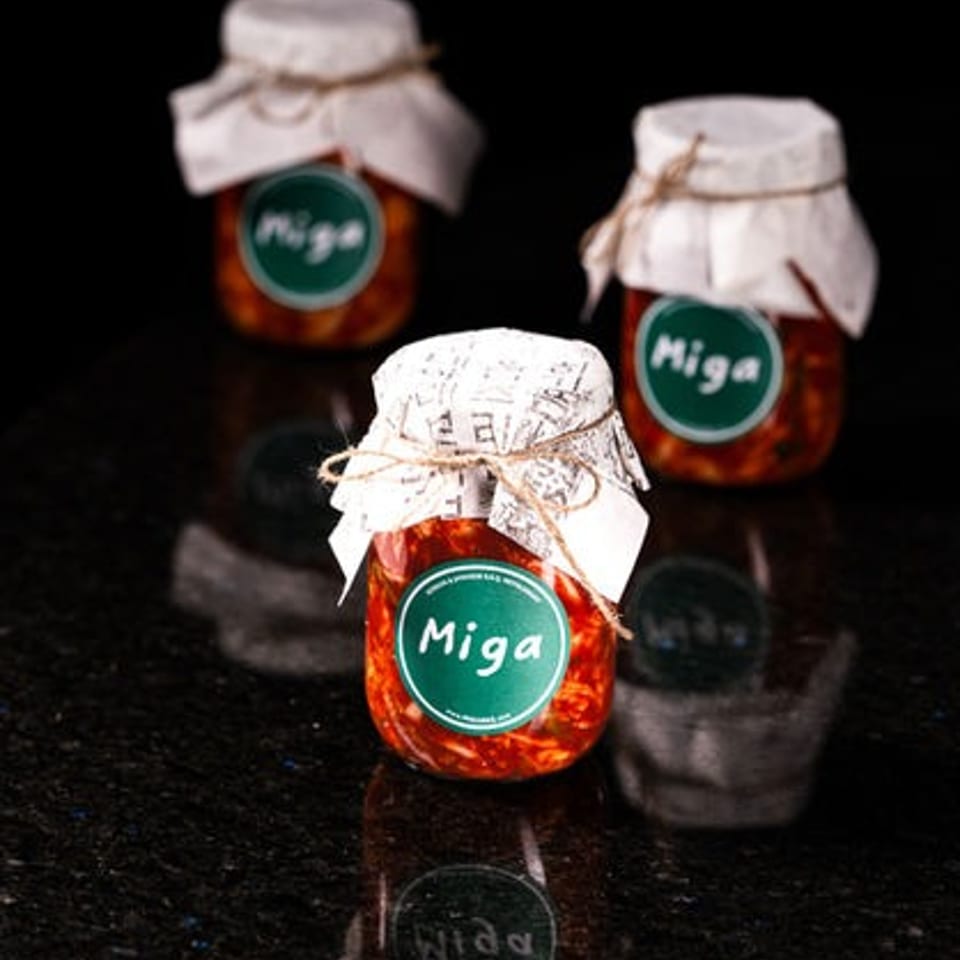 Three jars of red preserve labeled “Miga” with cloth-covered lids arranged on a reflective black surface.