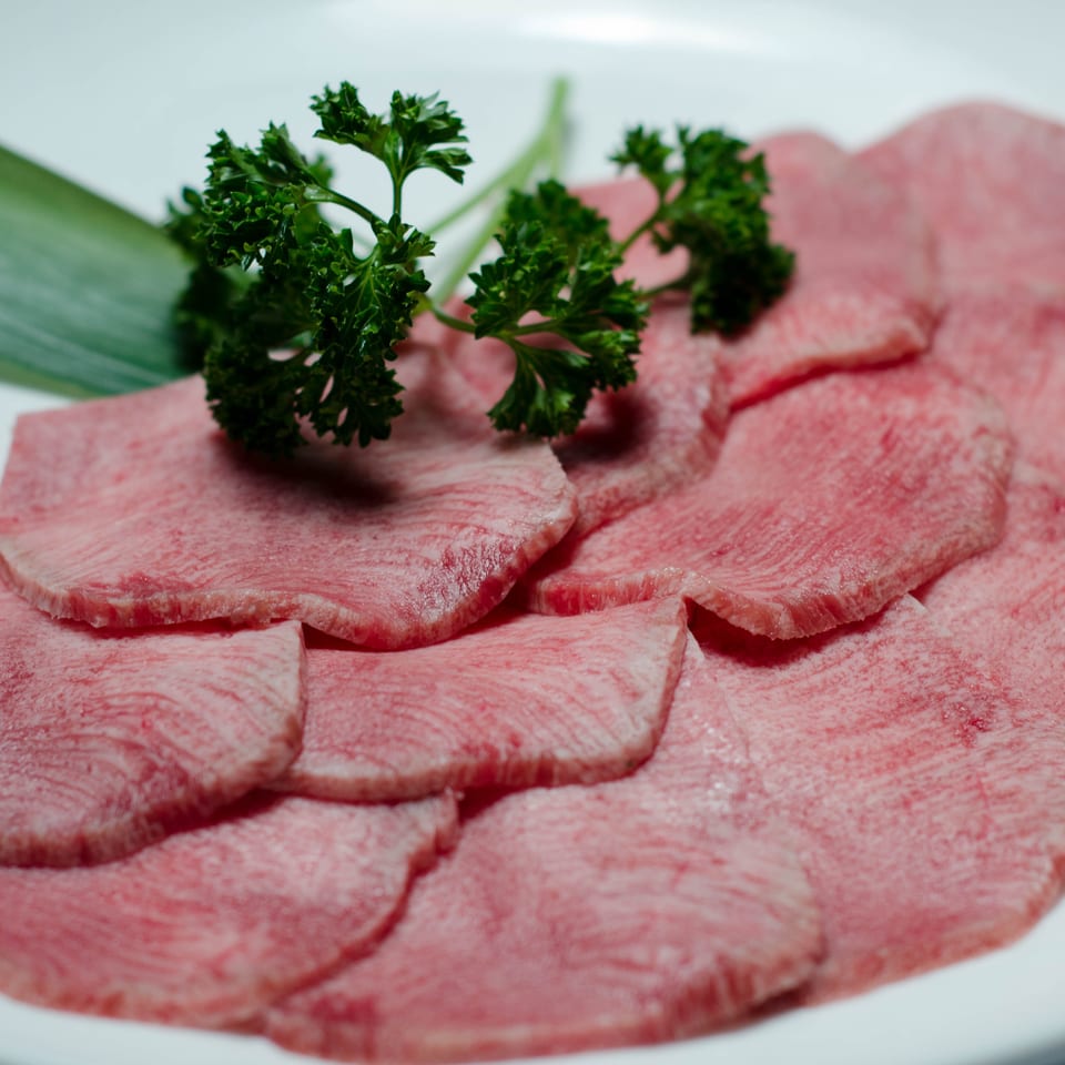 Thin slices of marbled raw beef arranged on a white plate with parsley garnish and a green leaf.