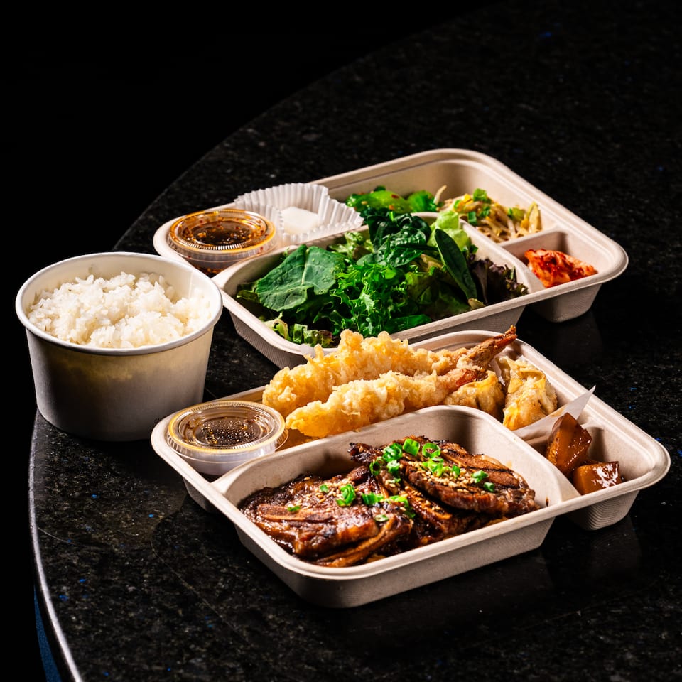 Japanese takeout spread with rice, tempura, glazed grilled fish, leafy greens, dipping sauces, and side dishes on a dark table.