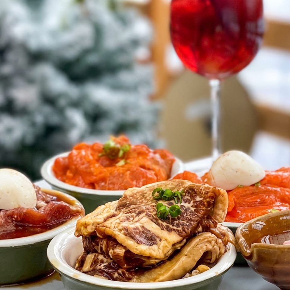 Assorted Korean dishes fill ceramic bowls, including sauced meats, dumplings, vegetables, and creamy garnishes beside a glass of red wine.