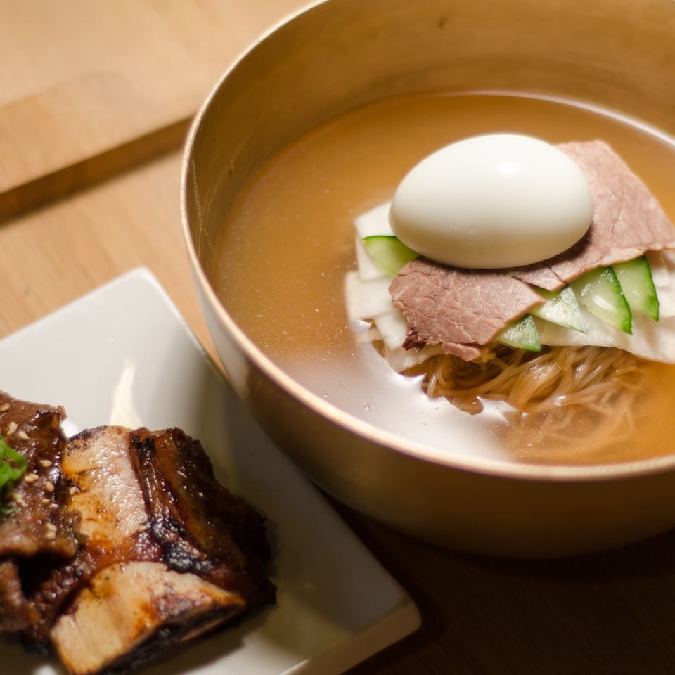 Bowl of clear broth noodles topped with sliced beef, cucumber, and a boiled egg beside grilled sesame-glazed meat.