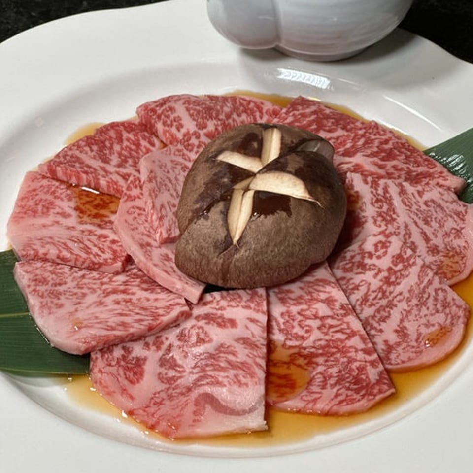 Marbled raw wagyu beef slices arranged around a cross-cut shiitake mushroom on a white plate with dipping sauce.