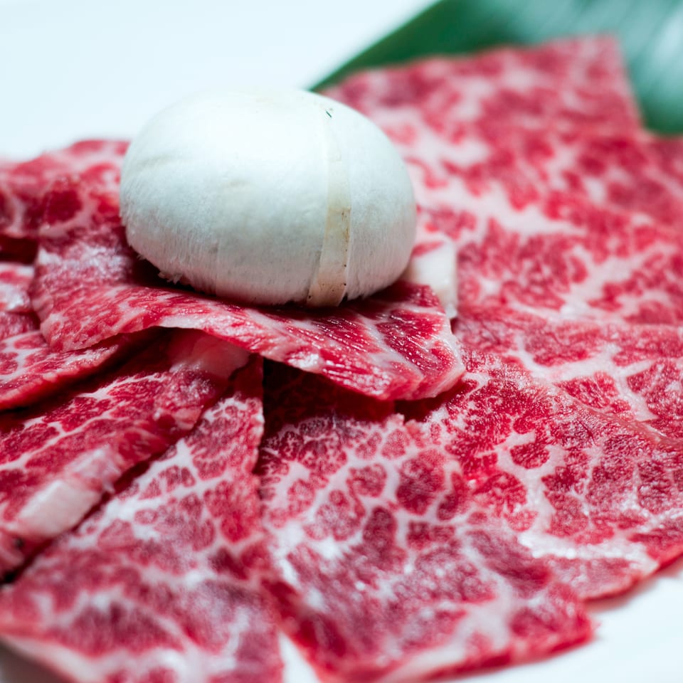 Thinly sliced, richly marbled raw beef arranged around a whole white mushroom on a plate.