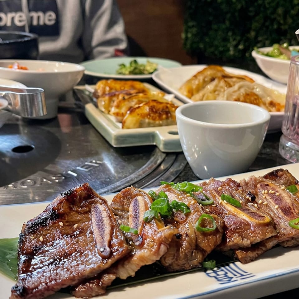 Grilled Korean-style ribs garnished with scallions and sesame seeds sit alongside dumplings, side dishes, and a pink drink on a restaurant table.