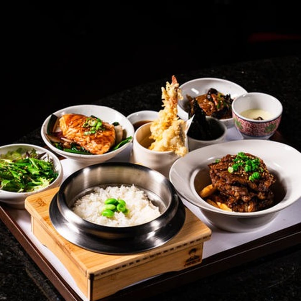 Korean-style meal spread with rice, grilled meat, fried shrimp, vegetables, side dishes, and tea on a wooden serving board.